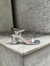 Sandalias plateadas con tiras finas, punta cuadrada, talón abierto y hebilla ajustable en el tobillo. Taco bajo y ancho de 7,5 cm.