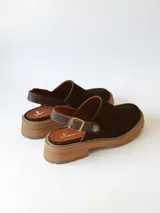 Zueco de cuero gamuzado color chocolate, con diseño de punta cerrada y tira ajustable con hebilla metálica en el talón. Presenta una suela de plataforma dentada en tono marrón claro.