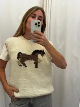 Chaleco tejido de punto color marfil, confeccionado en 100% lana merino, con cuello redondo y diseño frontal bordado artesanalmente que representa un caballo en tonos marrones y negros.