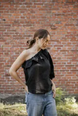 Blusa asimétrica de lino color negro, con un hombro al descubierto y detalle de volado en el escote. Presenta manga larga abullonada y corte recto.