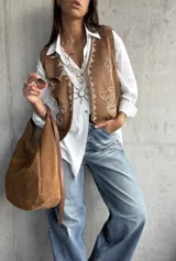 Chaleco de gamuza color camel con bordados en hilo blanco y detalles de tachas metálicas. Presenta un diseño abierto con terminaciones redondeadas en el frente.