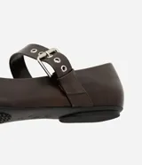 Zapatilla tipo bailarina color marrón oscuro, confeccionada en poliuretano con acabado mate. Presenta punta redondeada, suela plana y dos tiras ajustables con hebillas metálicas plateadas y ojales decorativos sobre el empeine.
