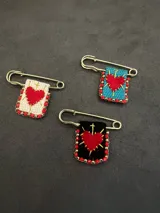 Pin de gancho metálico con dije rectangular bordado que presenta un corazón rojo central atravesado por espadas, decorado con cuentas rojas y doradas en el borde.