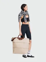 Bolso de viaje de la línea Le Pliage Original de Longchamp, diseñado en formato ampliable. Presenta un cuerpo de nylon en tono beige claro con detalles de cuero marrón en las asas y la solapa frontal con cierre de botón a presión. Incluye una correa ajustable y extraíble para llevar al hombro o cruzado.
