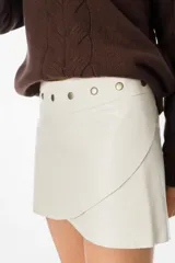 Mini falda de cuero sintético en color crema, con diseño cruzado tipo envolvente y detalle de tachas metálicas circulares en la cintura.