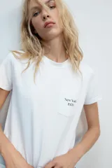Camiseta blanca de algodón con cuello redondo, manga corta y bolsillo en el pecho que incluye un texto impreso a contraste.