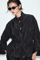 Cazadora satinada de color negro, con cuello subido, bolsillos con solapa en el pecho y cordón ajustable en la cintura. Cierre frontal con botones metálicos y mangas anchas.