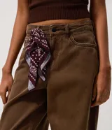 Pantalón de jean color marrón con corte baggy holgado, tiro medio y cierre frontal con cremallera y botón. Incluye un pañuelo estampado decorativo en la cintura.