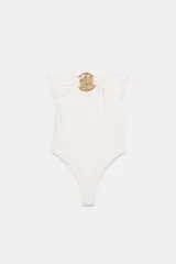 Bañador enterizo tipo bandeau color crema, con escote recto drapeado y detalle de pieza metálica circular dorada en el centro del pecho. Cuenta con forro interior.