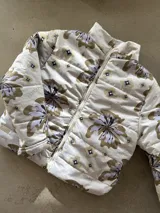 Campera puffer color beige con estampado floral y de logotipos en tonos marrones y grises, cuello alto y cierre frontal con cremallera.