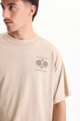 Remera beige de manga corta y corte oversize, con estampa en la espalda con temática de tenis y texto descriptivo.