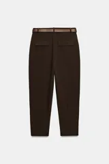 Pantalón tobillero de tiro medio, color beige, con pinzas delanteras y bolsillos frontales. Incluye un cinturón fino de cuero marrón con hebilla metálica dorada y cierre frontal con cremallera, botón interior y gancho.