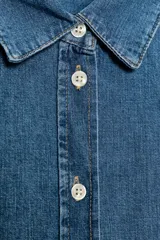 Camisa de denim azul de manga larga con cuello solapa y cierre frontal de botones. Presenta bajo redondeado.