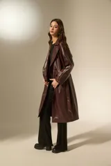 Trinchera de eco-cuero en color bordó, con diseño de doble botonadura, solapas amplias y cinturón ajustable en la cintura. Presenta mangas largas con detalles en los puños y bolsillos laterales.