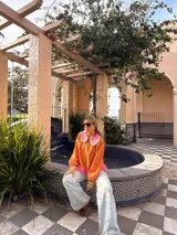 Camisa de manga larga color naranja con cuello grande en contraste color rosa y detalle de volados en el borde. Presenta un diseño con botones frontales y un detalle de tela rosa en la parte inferior.