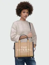 Cartera tipo tote bag Marc Jacobs modelo The Medium Leather Tote Bag, color marrón tostado con detalles en negro, confeccionada en cuero con las inscripciones "THE TOTE BAG" y "MARC JACOBS" estampadas en el frente. Cuenta con asas superiores y correa ajustable y desmontable para el hombro.