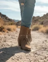 Bota de caña alta estilo western confeccionada en cuero vacuno color marrón oscuro. Presenta un diseño versátil con estribo desmontable y detalle de flecos con tachas metálicas en la base. Cuenta con taco cuadrado de madera y punta fina.