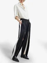 Pantalón negro de silueta relajada con pliegues frontales, cintura elástica y cierres metálicos largos en las piernas que revelan una apertura regulable y paneles laterales blancos.