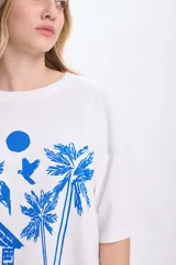 Remera blanca de corte amplio con estampado de palmeras, cabaña y aves en color azul.