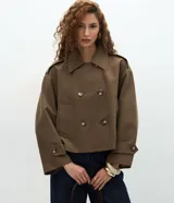 Trench coat corto de lona estructurada en color marrón, con diseño de abotonado cruzado, cuello clásico, mangas largas con trabillas en los puños y bolsillos frontales.