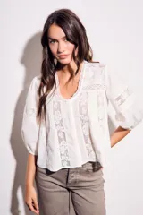 Blusa blanca de algodón peinado, corte oversize, con escote en V. Presenta detalles de encaje calado y bordados verticales en el frente, y mangas abullonadas de tres cuartos.
