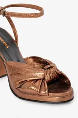Sandalia de noche color bronce metalizado con taco alto y plataforma. Presenta una capellada con nudo fruncido y tira fina ajustable en el tobillo con hebilla.