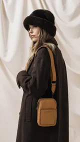 Bandolera pequeña de diseño compacto en color mostaza, confeccionada en material sintético con textura granulada. Presenta un bolsillo frontal con el logo de la marca impreso, cierre superior y correa ancha ajustable y desmontable.