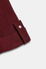 Camisa negra de textura arrugada, con cuello solapa, escote en V y cierre frontal de botones. Cuenta con mangas cortas terminadas en vuelta con trabilla y botón.