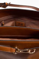 Cartera de hombro color tostado, fabricada en tejido revestido, con correa ajustable y un divisor interior que crea dos compartimentos, además de un compartimento interior con cierre. Presenta una trabilla decorativa con mosquetón metálico dorado en la parte delantera y una etiqueta extraíble.