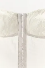 Corsé blanco de escote corazón y hombros descubiertos, confeccionado con copas de encaje y cuerpo de tejido de malla transparente con varillas interiores y cierre frontal de ganchos metálicos.