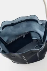 Bolso de hombro tipo saco confeccionado en piel azul marino, con detalle de pespuntes blancos verticales, asas finas de hombro en color blanco y cierre fruncido.