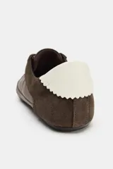 Zapatilla estilo blucher con diseño deportivo, confeccionada en una combinación de materiales textiles y gamuza en tono marrón, con detalle de contraste en color blanco en el talón y cierre mediante cordones.