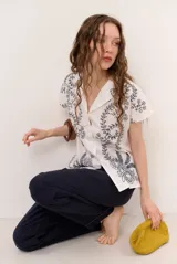 Camisa blanca de manga corta con cuello solapa, cierre frontal de botones y diseño bordado en hilo azul marino con motivos florales y vegetales.