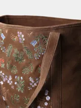 Bolso tipo tote bag confeccionado en cuero serraje color marrón, con diseño artesanal que incluye bordados florales coloridos en el panel frontal. Cuenta con asas de hombro y cierre superior.