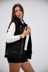 Chaleco negro de estilo aviador con interior y bordes de corderito sintético. Presenta cierre frontal metálico, detalles de tiras de cuero sintético y hebillas ajustables en el cuello y el ruedo.
