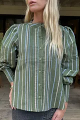 Camisa de algodón a rayas verticales en tonos verde oliva, celeste y crema. Presenta cuello redondo, cierre frontal con botones, diseño plisado en el frente y mangas largas abullonadas con puños ajustados.