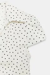 Camisa de cuello solapa y escote en pico, con mangas cortas y detalle de frunces centrales. Presenta cierre frontal con botones y estampado de lunares negros sobre fondo blanco.