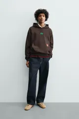 Sudadera con capucha de corte relajado, color marrón oscuro, con interior perchado. Presenta un bolsillo frontal tipo canguro, bordado con la palabra 'LOVE' en el pecho y un parche decorativo a contraste en el bolsillo. Posee puños y ruedo en rib.