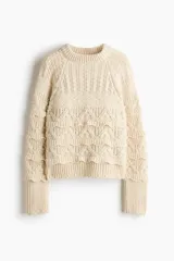 Buzo de punto color beige con textura, cuello redondo acanalado, mangas raglán y detalles de volados horizontales en el frente y las mangas. Presenta puños y ruedo acanalados con terminaciones festoneadas.