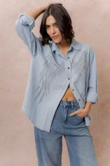 Camisa de denim liviano con calce relajado, cuello camisero clásico y cierre frontal con botones. Presenta una destacada aplicación de strass en el frente formando un diseño de alas.