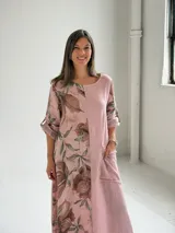 Vestido largo color rosa, con mangas cortas y diseño holgado, presenta un estampado floral en tonos marrones y verdes en un lateral, además de un bolsillo aplicado en la parte frontal y costuras visibles de estilo rústico.