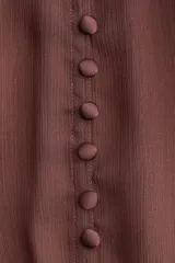 Blusa de tejido liviano color marrón oscuro con efecto arrugado. Presenta un corte holgado, cuello elevado, botones forrados en el frente y canesú fruncido de doble capa. Las mangas son globo a tres cuartos con puños ribeteados con cinta fina.