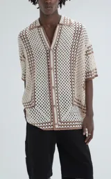 Camisa de manga corta con diseño de tejido abierto tipo crochet, corte oversize y cuello solapa. Presenta un patrón geométrico en tonos crema y marrón con cierre frontal de botones.