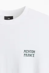 Remera blanca de punto de algodón, con cuello redondo y hombros caídos, corte holgado. Presenta un pequeño estampado en el pecho con el texto "MENTON FRANCE" en color oscuro.