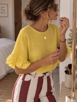 Blusa blanca de tela liviana y fresca, con mangas cortas abullonadas terminadas en volados y detalle de volado en el ruedo. Presenta escote redondo con pequeña abertura y botón en la parte posterior.