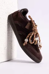 Zapatillas urbanas de gamuza color marrón, con diseño de cordones decorados con cuentas de piedra y detalles de líneas brillantes en los laterales. Presentan suela de goma texturizada al tono.