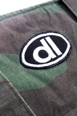 Bolso tote de tela con estampado de camuflaje en tonos verdes y marrones. Tiene asas de hombro y un logo ovalado con letras blancas en el centro.