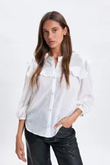 Camisa blanca de corte casual con cuello redondo, cierre frontal de botones y detalle de volados en los hombros. Presenta mangas tres cuartos con puños elásticos.