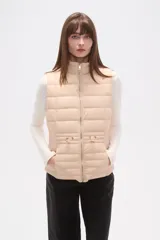 Chaleco acolchado color beige claro, sin mangas, con cuello alto y cierre frontal de cremallera dorada. Presenta detalles metálicos dorados en los bolsillos delanteros.