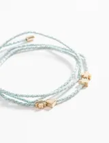 Pulsera de cordón trenzado color celeste con hilo plateado, con dijes metálicos dorados en forma de corazón, trébol de cuatro hojas y cruz.